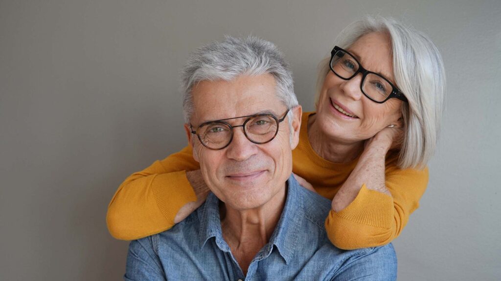 A senior couple with grey hair and glasses smiling warmly; the woman wears a mustard yellow sweater and leans her chin on the man's shoulder while he wears a blue denim shirt, both posed against a neutral grey background.