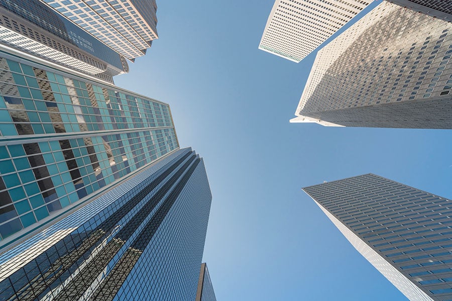 A low-angle view looking straight up at several modern glass skyscrapers towering against a clear blue sky.