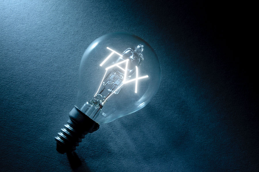 A clear glass lightbulb resting on a dark, textured surface bathed in blue light, with its internal glowing filament shaped to spell the word "TAX".