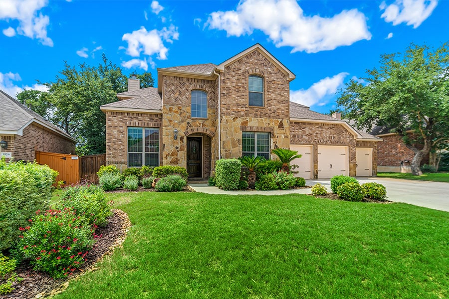 A large two-story brick and stone house with a three-car garage, featuring a vibrant green lawn and manicured landscaping under a bright blue sky with white clouds.
