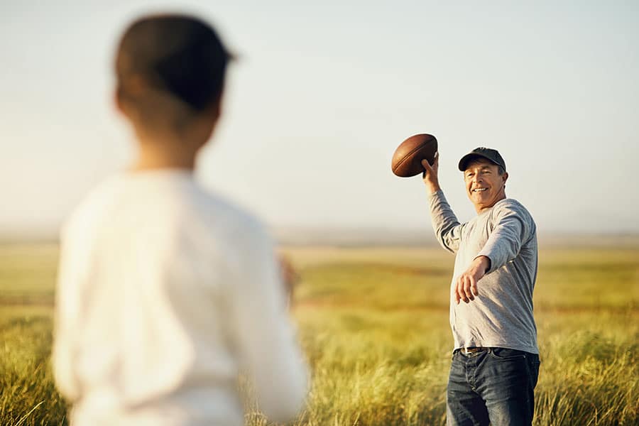 A smiling man in a baseball cap stands in a grassy field about to throw a football to a young boy, whose back is to the camera in the foreground.