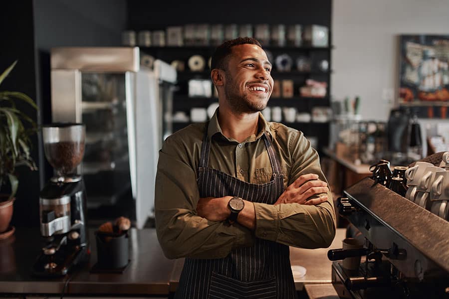 A smiling young man in a striped apron stands with his arms crossed in a coffee shop, looking off-camera with a proud and happy expression.