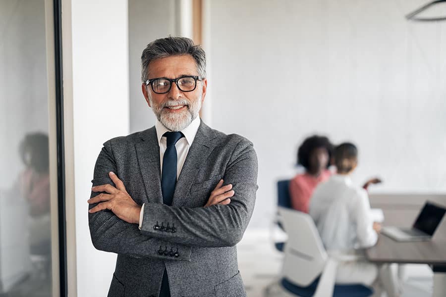 A confident, middle-aged professional man with grey hair, a beard, and glasses stands with his arms crossed in a modern office. He is wearing a grey suit jacket and a dark tie. In the blurred background, two colleagues are seated at a desk with a laptop, engaged in a discussion.