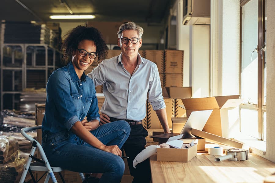 Two smiling colleagues, a woman seated and a man standing, looking directly at the camera in a sunlit stockroom next to a wooden counter with an open laptop, packing tape, and cardboard shipping boxes.