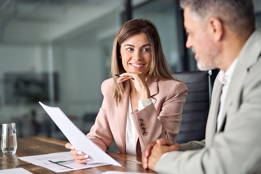 A professional woman in a mauve blazer smiling as she listens to a man in a grey suit during a consultation. She holds a pen and a set of financial documents at a wooden conference table in a modern office setting.