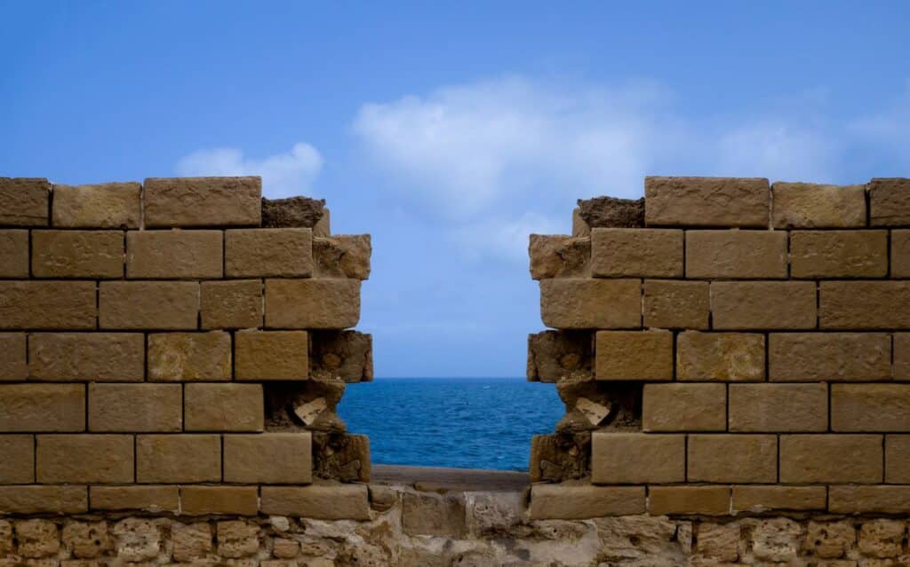 A stone wall with a gap overlooks the blue Mediterranean Sea under a clear sky.