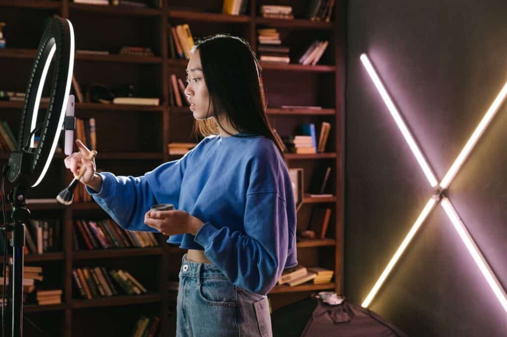 A woman who is a content creator in a blue sweater applying makeup in front of a ring light, set indoors.
